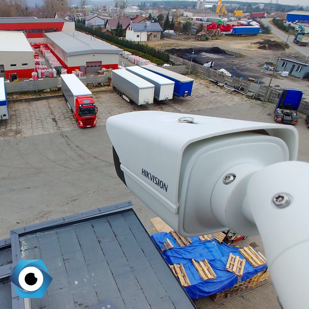 Kamera przemysłowa Hikvision zamontowana na dachu, monitorująca plac manewrowy z zaparkowanymi samochodami ciężarowymi i naczepami.