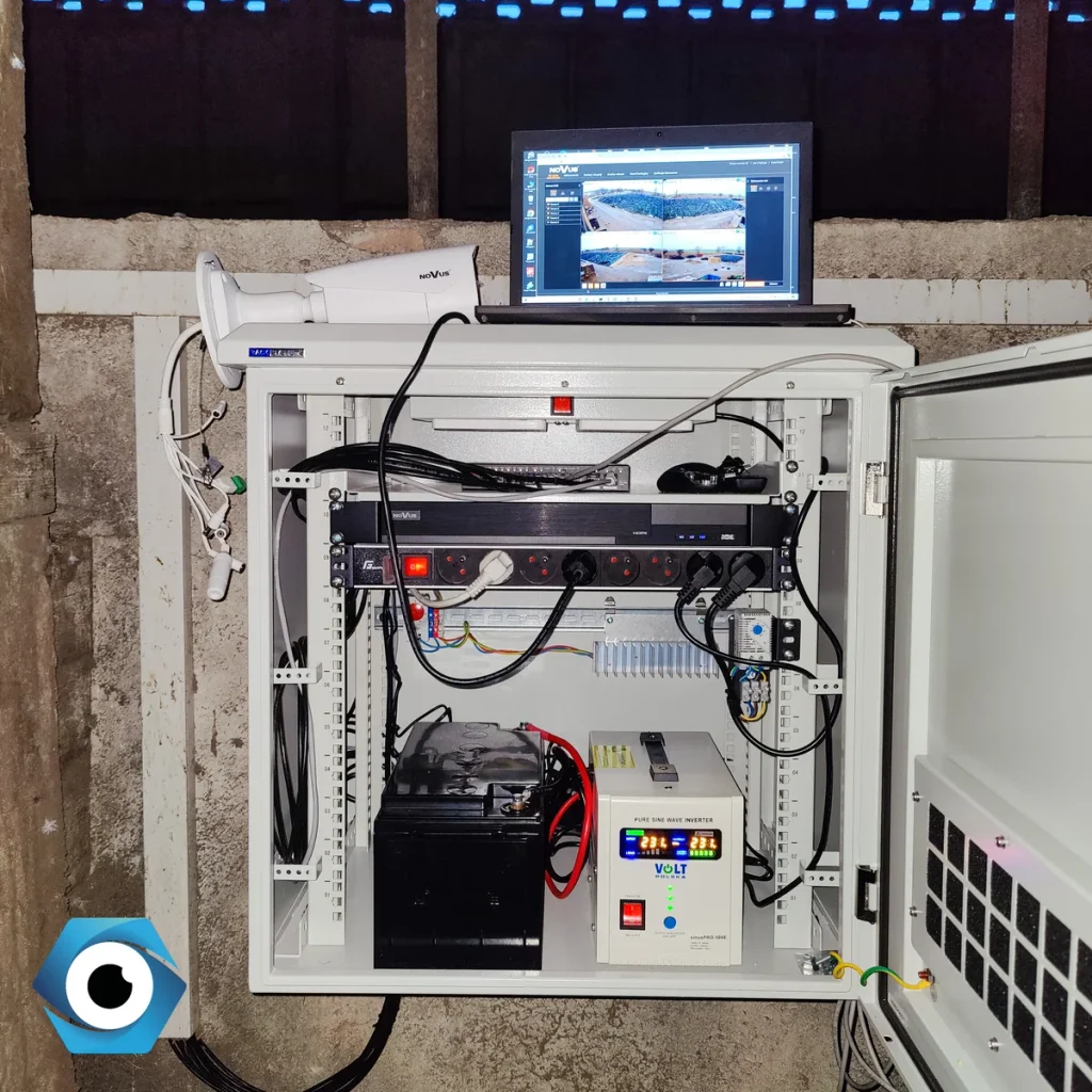 Wnętrze profesjonalnej szafy rackowej z osprzętem do monitoringu, zawierającej akumulator, falownik oraz laptop wyświetlający podgląd z kamer na gospodarstwie.
